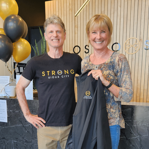 Man & woman smiling at OsteoStrong Sioux City holding branded apparel inside the wellness center.