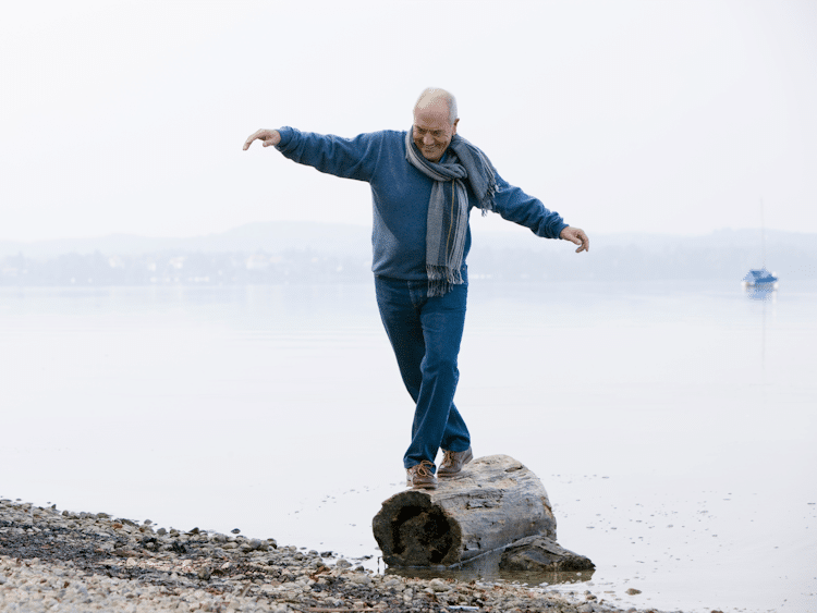 Elderly man balancing on a log by the lakeside with arms stretched out.